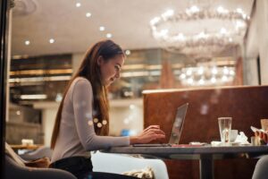 A young woman with long hair sits at a café table, focused on her laptop while typing. The warm indoor lighting and a large, glowing chandelier in the background create a cozy, modern atmosphere.