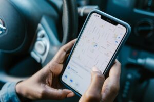 A person's hands hold a smartphone displaying a navigation map while sitting behind the steering wheel of a car. The bright screen shows a detailed street layout with various location markers and a search bar for finding destinations.