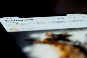 This image shows a close-up, angled view of a smartphone screen displaying the Instagram app interface. The focus is sharpest on the "Instagram" logo and the top icons, while the rest of the feed fades into a soft blur.