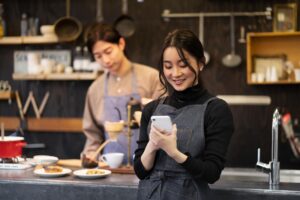 A woman in a dark denim apron smiles while looking at her smartphone in a cozy, rustic kitchen setting. In the blurred background, a man in a matching apron carefully prepares pour-over coffee near a display of kitchenware and small plates of food.