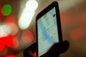 A person holds a smartphone displaying a digital map of New York City against a background of soft, colorful bokeh lights. The image captures a modern urban moment, emphasizing the glow of the screen and the vibrant nighttime atmosphere.