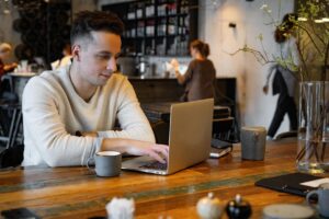 A young man in a white sweater sits at a rustic wooden table in a modern, industrial-style cafe while focused on his laptop. The candid scene captures a bustling background with soft natural lighting and other patrons, creating a quiet moment of productivity amidst a busy environment.