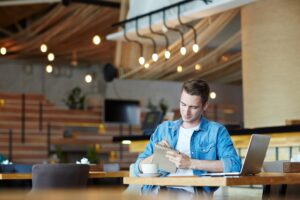 A young man sits at a wooden table in a modern cafe, focused on writing in a small notebook. A laptop, a cup of coffee, and warm overhead lighting create a productive and cozy atmosphere around him.