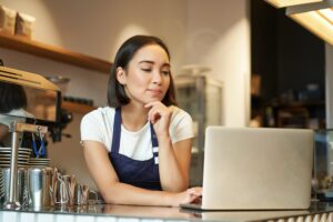 A barista wearing a navy blue apron stands behind a cafe counter while looking thoughtfully at a silver laptop. In the background, the shop is filled with warm lighting, stacked cups, and professional espresso equipment.