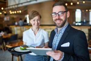 A bearded man in a blazer smiles while looking at a tablet in a modern restaurant setting. In the background, a server holds a tray with a burger on a charcoal bun, looking toward the man.