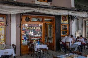 A traditional kebab restaurant storefront features a glass display filled with fresh tomatoes and peppers under a wooden-framed entrance. Outside, customers sit at small tables with white cloths, enjoying a meal in the shaded outdoor seating area.