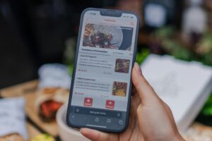A hand holds a smartphone displaying a digital menu for a restaurant called Yalinha, which features various combos and food items like esfihas. The background is softly blurred, showing glimpses of a dining setting with food and a white box on a table.