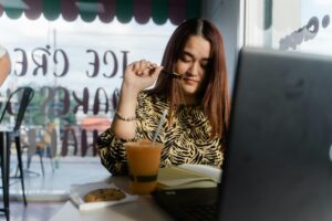 A young woman with reddish-brown hair sits at a cafe table, pensively holding a pen to her chin while looking at an open notebook. A laptop, a vibrant orange smoothie, and a chocolate chip cookie are spread out before her as she works near a window.