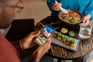 A person in a red sweater holds a smartphone to take a photo of a sushi roll and a bowl of ramen on a wooden table. Their dining companion sits across from them, holding a white ceramic spoon over the steaming bowl of noodles.