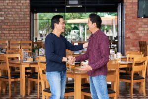 Two men are smiling and shaking hands inside a warm, casual restaurant with brick walls and wooden furniture. One man wears a dark blue long-sleeved shirt, while the other is dressed in a red and blue checkered button-down.