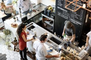 An overhead shot captures a busy, modern cafe with a barista in a beanie preparing drinks behind a marble counter. Customers wait at the counter and sit at nearby tables, surrounded by lush green plants and a large chalkboard menu.