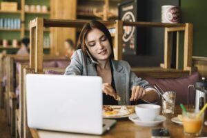 A woman in a grey blazer sits at a cafe table, multitasking by talking on her phone while eating breakfast in front of her laptop. The cozy setting features wooden furniture, a white coffee cup, and a plate of eggs and toast, suggesting a busy morning start.
