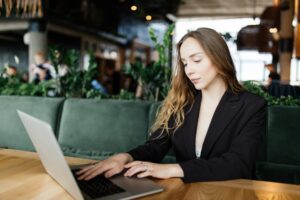A woman sits at a wooden table in a brightly lit cafe, focused on her work while typing on a silver laptop. She wears a professional black blazer and is positioned against a background of lush green plants and soft, ambient lighting.