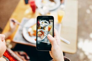 A person holds a smartphone over a wooden table to capture a top-down photo of their brunch, which includes eggs, toast, and orange juice. The background is softly blurred, focusing attention on the vivid display of the meal on the phone screen.