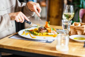 A person is shown using a knife and fork to cut into a plated meal featuring steak, asparagus, and a vibrant orange sauce. The table is elegantly set with a glass of white wine, a lit candle, and a basket of bread in a sunlit restaurant setting.