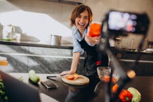 A cheerful woman in a black apron laughs while holding up a piece of sliced red pepper toward a camera on a tripod. The bright kitchen setting includes a wooden cutting board with fresh vegetables and a laptop used for filming her cooking vlog.