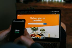 A person holds a smartphone displaying the Uber Eats logo in front of a laptop. The computer screen shows a Dutch food delivery website with the text "Tijd om eten te bestellen."