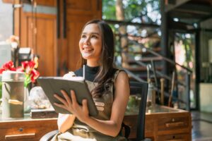 A smiling woman sits at a wooden desk in a sunlit, rustic room while holding a tablet and stylus. She is wearing an apron and looking upward with an inspired expression, surrounded by lush greenery and warm interior details.