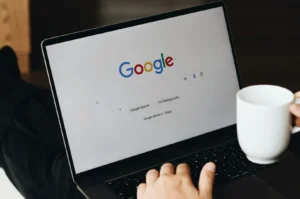 A laptop displays the Google search homepage. A person holds a white mug nearby, suggesting a relaxed, focused work or browsing environment.