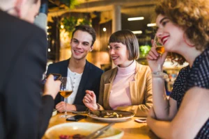 A group of four young adults is gathered around a table at a dimly lit, modern restaurant, smiling and engaged in conversation. One person in the foreground is holding a smartphone, while plates of food and glasses of wine are scattered across the wooden tabletop.