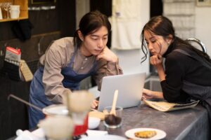 Two people wearing aprons lean over a counter in a café setting to look closely at a laptop screen. They appear focused and collaborative as they review information together in their workspace.