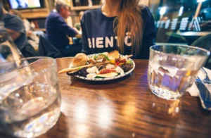 A vibrant plate of antipasto, featuring mozzarella, grilled vegetables, and crusty bread, sits on a wooden restaurant table between two glasses of water. In the soft-focus background, a person wearing a navy "BIENTÔT" sweatshirt prepares to enjoy the meal amidst the cozy hum of other diners.
