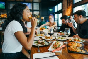 A smiling woman in a white t-shirt takes a bite of food while sitting at a long wooden table filled with various dishes and drinks. In the background, a diverse group of friends enjoys their meals and conversations in a lively, dimly lit restaurant setting.