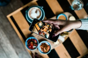 A top-down view shows a person holding a smartphone to take a photo of a breakfast spread on a wooden table. The phone screen displays a clear shot of the coffee, small bowls of food, and a main dish arranged below.