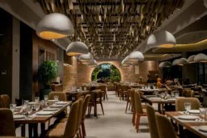 This restaurant interior features rows of elegant wooden tables set with crystal glassware and round, white pendant lights that resemble organic mushrooms. A dramatic ceiling installation of suspended golden panels leads the eye toward a circular brick entryway framed by lush greenery.