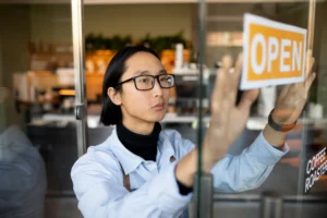 In the image, a person wearing glasses and an apron carefully places a yellow "OPEN" sign on a glass door. This individual appears focused while preparing to welcome customers into what looks like a modern coffee roastery.