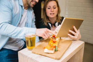 A man and a woman sit at a wooden table while looking at a gold tablet held by the woman. In front of them on the table is a long, flatbread-style pizza on a wooden board next to a glass of beer.