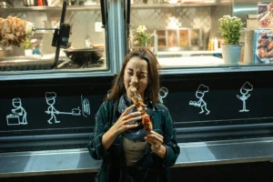 A woman with long brown hair stands in front of a food truck, closing her eyes as she takes a bite from a large grilled food skewer. The food truck behind her features stick-figure chef drawings on its side and reveals a brightly lit kitchen interior with a vertical rotisserie.