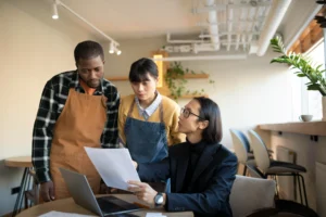 Two shop or café owners wearing aprons look on anxiously as they review a document with a consultant or lawyer in a modern café setting. The professional, seated at a laptop, points to the paper while explaining its contents to the standing couple.