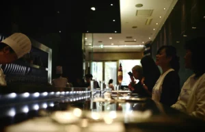A chef wearing a white toque works behind a sleek, dimly lit counter while patrons sit and wait for their orders. The modern interior features reflective surfaces, accent lighting along the bar, and tall glasses set out for the guests.