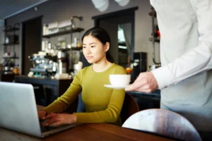 A young woman sits at a wooden table in a modern cafe, focused on her laptop screen. A server in a grey apron stands beside her, carefully handing over a small white cup and saucer.