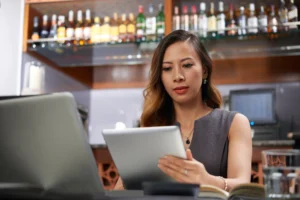A woman sits at a table in a bar or restaurant, focused on the digital tablet she is holding in her hands. A laptop and open notebook are also on the table, while shelves stocked with various liquor bottles visible in the background suggest a professional setting.