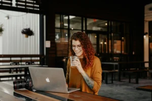 A smiling woman with curly reddish-brown hair sits at an outdoor wooden table, drinking an iced beverage while looking at her laptop. A professional camera rests on the table next to her laptop against a blurred cafe background.