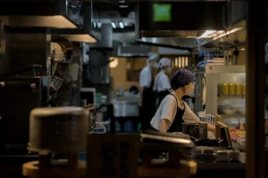 A focused kitchen staff works in a dimly lit, narrow restaurant kitchen filled with stainless steel equipment and stacked bowls. The workers wear masks and traditional head coverings while preparing food under the warm glow of overhead lights.