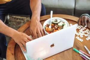 An over-the-shoulder view shows a person typing on an open silver MacBook at a wooden coffee table. Nearby sits a bowl of food with sliced avocado, scattered white letter tiles, and several colored pens.