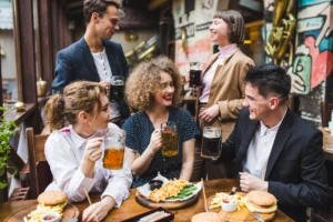 A group of five friends laughs and talks while gathered around a wooden table at a restaurant. They are holding glass mugs of beer and enjoying a meal of burgers, fries, and waffles.