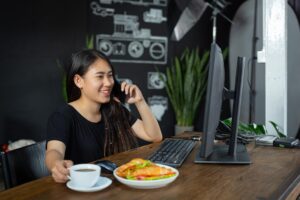 A smiling woman sits at a wooden desk with a computer, talking on her smartphone while reaching for a cup of coffee. On the desk, a plate with two croissants filled with greens and tomatoes sits next to her keyboard.