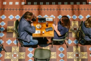 An overhead, birds-eye view shows a man and a woman engaged in conversation at a wooden cafe table. They sit on green chairs on a vibrant, intricately patterned tiled floor featuring geometric shapes in red, blue, and yellow.