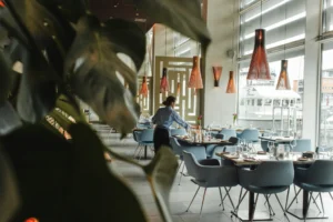 A server in a light blue shirt sets a table inside a modern, sunlit restaurant featuring floor-to-ceiling windows and distinctive ribbed pendant lights. Large monstera leaves frame the foreground, while a white boat is visible through the glass in the background.