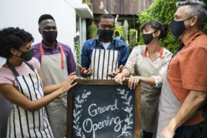 In the image, a diverse group of five people wearing face masks and aprons gathers around a chalkboard sign that reads "Grand Opening." They are posing outdoors in a lush, greenery-filled space, appearing cheerful as they celebrate the start of their new business venture.
