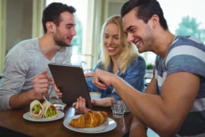 Three young friends smile while sitting together at a cafe table and looking at a digital tablet. In front of them are plates with a croissant and wraps, along with glasses of water and coffee.
