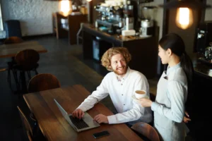 A man with curly hair sits at a wooden table, smiling as a barista serves him a cup of coffee while he works on his laptop. The cafe features a warm, industrial aesthetic with soft lighting and a professional espresso machine visible in the background.