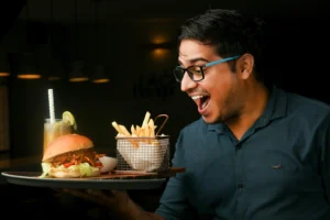 A smiling man wearing glasses looks excitedly with an open mouth at a tray of food he is holding. The tray contains a burger, a wire basket of french fries, and a tall drink with a lime slice garnish.