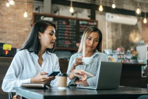 Two women sit together at a wooden table inside a cozy coffee shop while collaborating over a laptop. The woman on the left holds a smartphone and gestures toward the laptop screen, while the woman on the right looks at the screen while holding a white coffee cup.