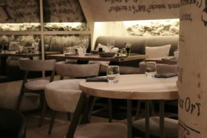 This image showcases an upscale restaurant interior featuring light wood tables paired with soft, cushioned seating. Warm, ambient lighting illuminates textured walls adorned with elegant script, creating a sophisticated and cozy dining atmosphere.