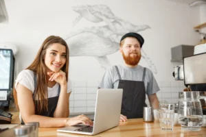 A woman in a black apron smiles while working on a laptop at a bright, modern cafe counter. Behind her, a bearded man in matching attire stands near an espresso machine, with a large whale mural decorating the wall in the background.